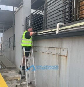 TM Waterproofing expert working on the side of a metal carport structure near pipes, applying waterproofing treatment.