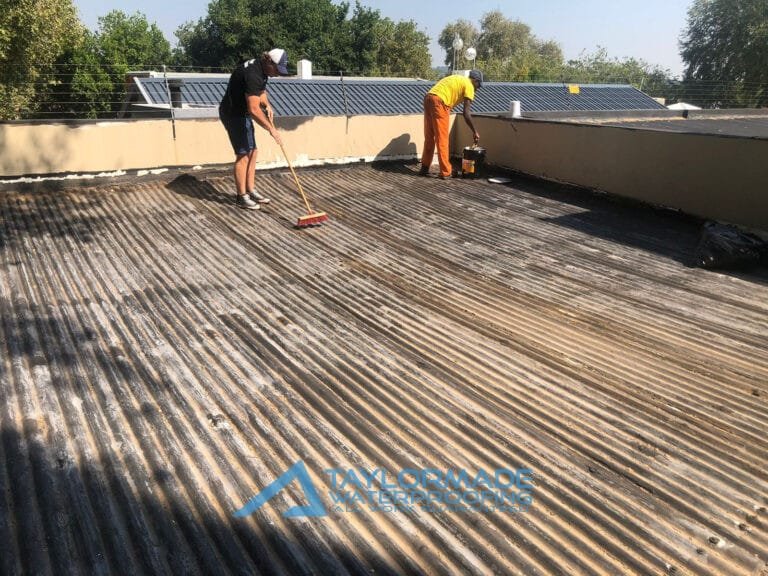 TM Waterproofing painting the side wall of a corrugated roof while expert sweeps surface in preparation for protective coating.