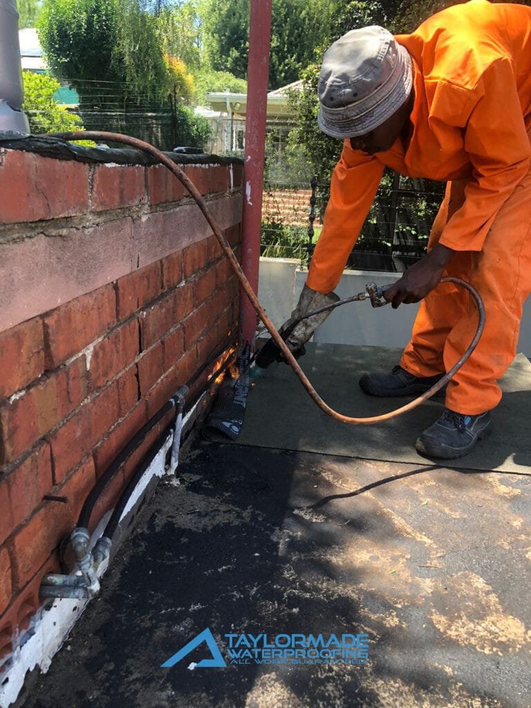 TM Waterproofing applying torch‑on waterproofing membrane on flat roof while wearing safety gear.