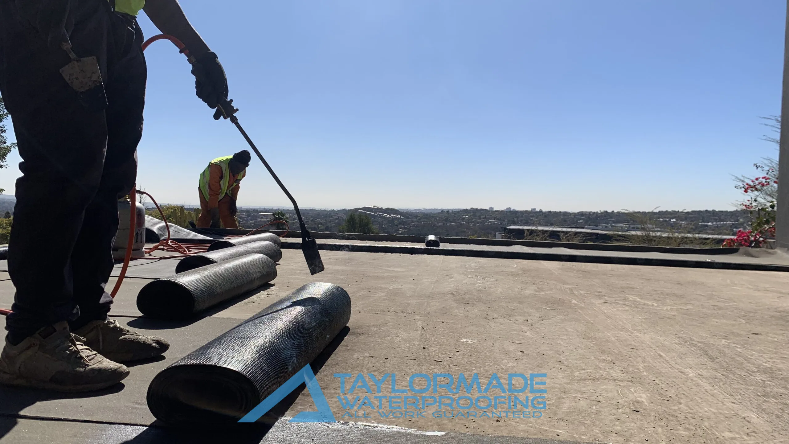 TM Waterproofing applying a torch‑on waterproofing membrane; close view of one technician holding the torch during application.