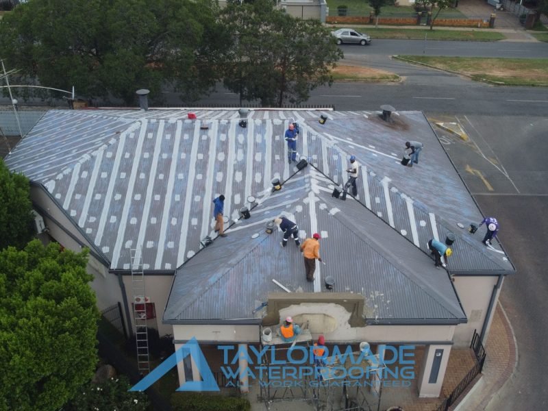Front drone view showing TM Waterproofing applying protective paint on a corrugated roof with construction work ongoing at the front entrance.