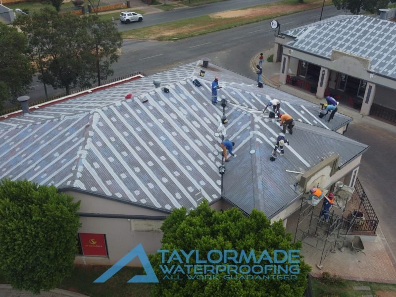 Drone view of TM technicians applying protective paint on a corrugated roof while construction continues at the front entrance.