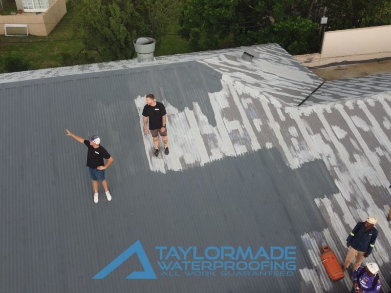 Drone view of TM workers and experts on a corrugated roof overseeing the application of membranes and protective paint.