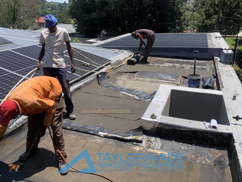 TM Waterproofing on a flat slab roof around a skylight in Parkhurst applying membranes, one holds pipes supports, one uses a torch-on membrane tool, the third installs board substrate.