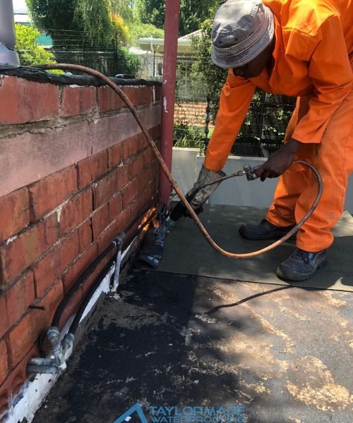 TM Waterproofing applying torch‑on waterproofing membrane on flat roof while wearing safety gear.